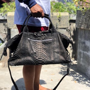 May include: A black handbag with a snakeskin pattern. The bag has a zipper closure and two handles, as well as a long shoulder strap. The bag is held up by a person.