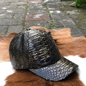 May include: A baseball cap featuring a textured, scale-like pattern in black and metallic gold. The cap has a structured crown and a curved brim, with the material displaying a distinct, repeating pattern across the entire surface of the headwear.