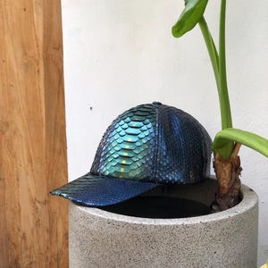 Könnte beinhalten: Eine Baseballkappe mit einem strukturierten, irisierenden Schuppenmuster, das zwischen tiefblauen, türkisfarbenen und grünen Farbtönen wechselt. Die Oberfläche hat ein reflektierendes, metallisches Finish mit kleinen, goldenen Akzenten am Mittelteil.