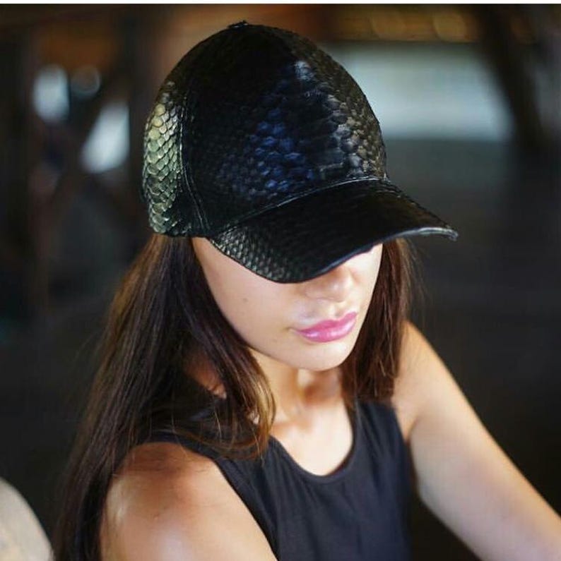 May include: A black baseball cap featuring a textured, scale-like pattern across the entire surface. The cap has a structured crown and a curved brim, constructed from a material that mimics the appearance of snakeskin with a glossy finish.