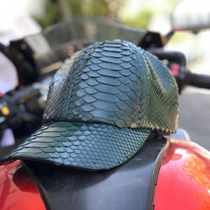 May include: A dark green baseball cap with a textured, scale-like pattern. The cap is made of a material that resembles snakeskin. The cap is resting on a red and black surface.
