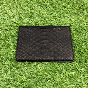 May include: A black leather card holder with a textured, snake-like pattern. The wallet is rectangular and appears to be made of high-quality leather. The card holder is set against a backdrop of green grass.