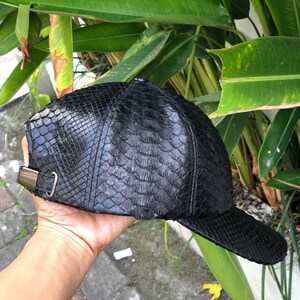 May include: A black baseball cap featuring a textured, scale-like pattern across the entire surface. The cap includes a curved brim and an adjustable metal buckle strap on the side, providing a secure fit. The material has a glossy finish.