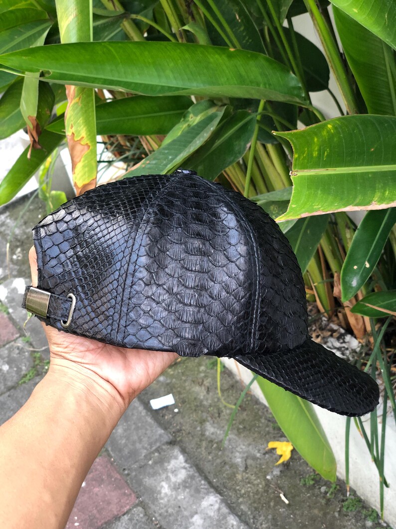 May include: A black baseball cap featuring a textured, scale-like pattern across the entire surface. The cap includes a curved brim and an adjustable metal buckle strap on the side, providing a secure fit. The material has a glossy finish.