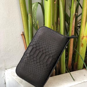 K&ouml;nnte beinhalten: Eine dunkelgraue, rechteckige Clutch mit Schlangenmuster. Die Clutch hat eine kleine Handschlaufe und einen Rei&szlig;verschluss. Der Hintergrund zeigt gr&uuml;ne Pflanzen und eine wei&szlig;e Wand.