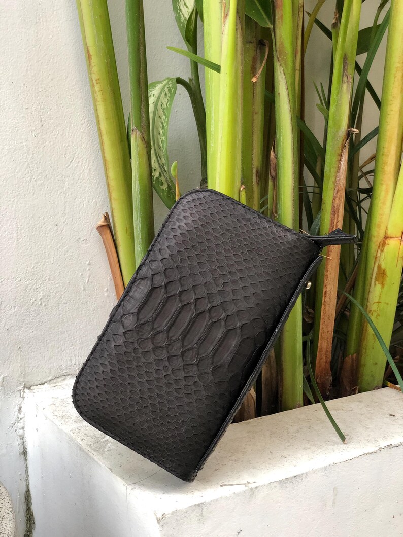K&ouml;nnte beinhalten: Eine dunkelgraue, rechteckige Clutch mit Schlangenmuster. Die Clutch hat eine kleine Handschlaufe und einen Rei&szlig;verschluss. Der Hintergrund zeigt gr&uuml;ne Pflanzen und eine wei&szlig;e Wand.