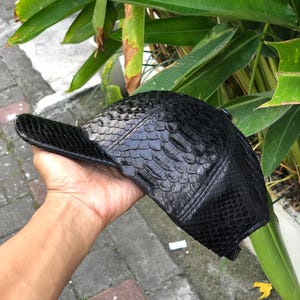 May include: A black baseball cap featuring a textured, scale-like pattern across the entire exterior surface. The cap has a structured crown and a curved brim, with visible stitching details along the panels and a button at the top centre.
