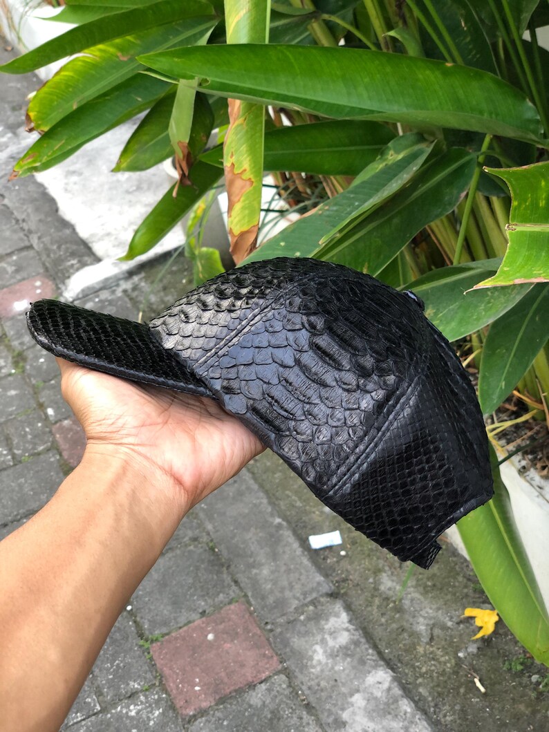 May include: A black baseball cap featuring a textured, scale-like pattern across the entire exterior surface. The cap has a structured crown and a curved brim, with visible stitching details along the panels and a button at the top centre.