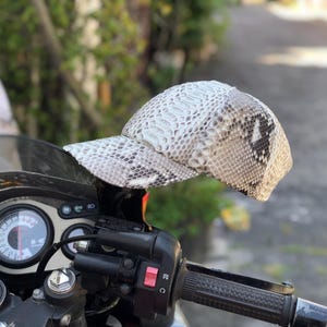 Può includere: Un cappellino da baseball con un motivo a pelle di serpente nei toni del bianco, grigio e nero. Il cappellino è esposto su una moto, mettendo in risalto il suo design e la sua texture unici. La visiera è curva e lo stile generale è casual.