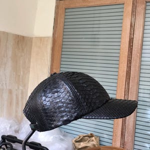 May include: A black baseball cap featuring a textured, scale-like pattern across the entire exterior surface. The cap has a rounded crown and a stiff, horizontal brim extending from the front, showcasing a uniform dark finish throughout.