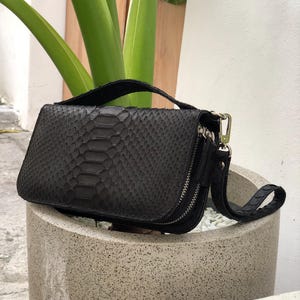 Puede incluir: Un bolso negro rectangular con un diseño de piel de serpiente. El bolso tiene cierre de cremallera, una correa de muñeca y un asa superior. El bolso está colocado sobre una maceta de hormigón, con plantas verdes al fondo.