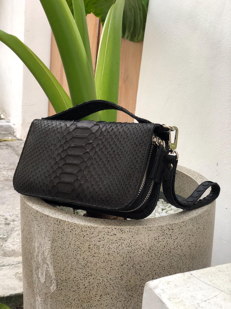 K&ouml;nnte beinhalten: Eine schwarze, rechteckige Handtasche mit Schlangenmuster. Die Tasche hat einen Rei&szlig;verschluss, eine Handschlaufe und einen oberen Griff. Die Tasche liegt auf einem Betonpflanzgef&auml;&szlig;, im Hintergrund sind gr&uuml;ne Pflanzen zu sehen.