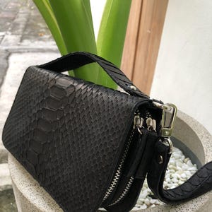 K&ouml;nnte beinhalten: Schwarze Schlangenleder-Clutch mit Henkel und abnehmbarem Riemen. Die Clutch hat zwei Rei&szlig;verschlussf&auml;cher und einen silberfarbenen Verschluss. Die Tasche steht in einem wei&szlig;en Steintopf mit gr&uuml;nen Pflanzen im Hintergrund.