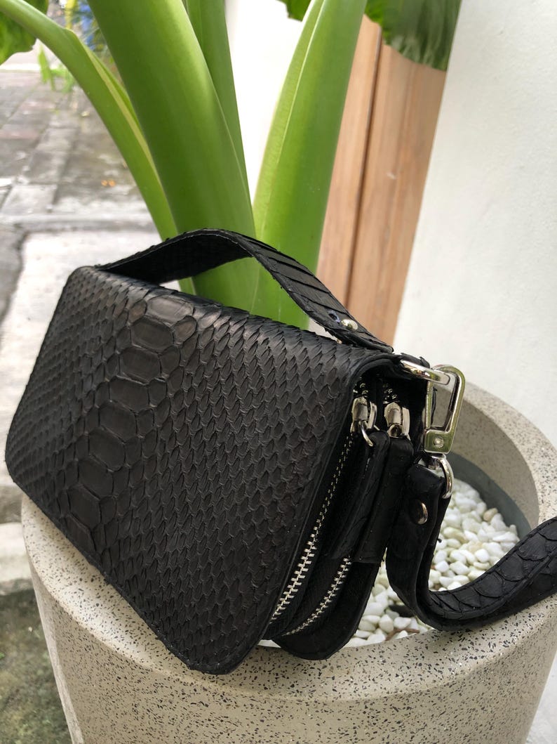 K&ouml;nnte beinhalten: Schwarze Schlangenleder-Clutch mit Henkel und abnehmbarem Riemen. Die Clutch hat zwei Rei&szlig;verschlussf&auml;cher und einen silberfarbenen Verschluss. Die Tasche steht in einem wei&szlig;en Steintopf mit gr&uuml;nen Pflanzen im Hintergrund.