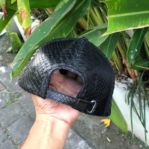 May include: A black baseball cap featuring a textured snakeskin pattern across the entire exterior. The cap includes an adjustable strap at the back with a metallic silver buckle closure, providing a secure fit for the wearer's head.