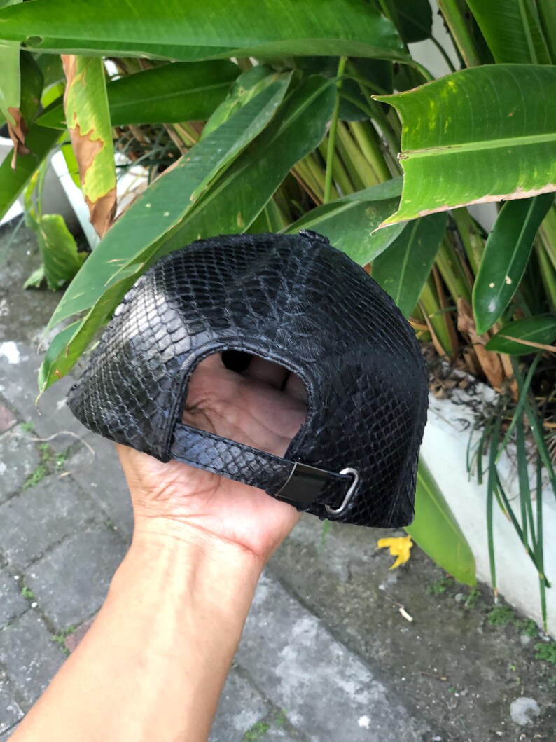 May include: A black baseball cap featuring a textured snakeskin pattern across the entire exterior. The cap includes an adjustable strap at the back with a metallic silver buckle closure, providing a secure fit for the wearer's head.