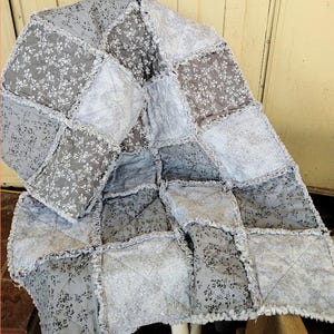 May include: A handmade patchwork quilt and pillow set. The quilt features a variety of gray fabrics with a floral pattern, arranged in a grid. The edges are ruffled. The pillow matches the quilt's design. A cozy, decorative bedding set.