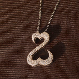 May include: A silver necklace with a double heart pendant encrusted with small, sparkling stones. The pendant is suspended from a delicate chain, creating a romantic and elegant look. The necklace is displayed against a brown textured background.