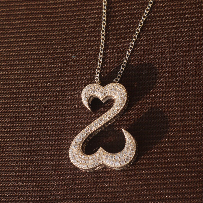 May include: A silver necklace with a double heart pendant encrusted with small, sparkling stones. The pendant is suspended from a delicate chain, creating a romantic and elegant look. The necklace is displayed against a brown textured background.