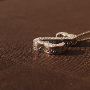 May include: A silver pendant necklace with a cursive letter "S" design. The pendant is encrusted with small, sparkling stones. The necklace chain is delicate and silver. The pendant is approximately 1 inch in length.