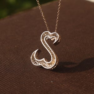 May include: A silver-toned pendant necklace featuring the letter "S" in a heart-shaped design. The pendant has a filigree pattern and hangs from a delicate chain. The necklace is displayed on a brown surface.