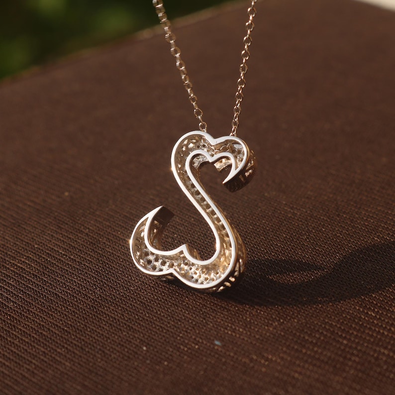 May include: A silver-toned pendant necklace featuring the letter "S" in a heart-shaped design. The pendant has a filigree pattern and hangs from a delicate chain. The necklace is displayed on a brown surface.