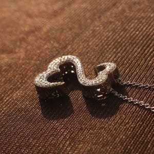 May include: A silver-colored pendant necklace featuring a double heart design. The pendant is encrusted with small, sparkling stones. The necklace chain is delicate and silver, resting on a brown textured surface.