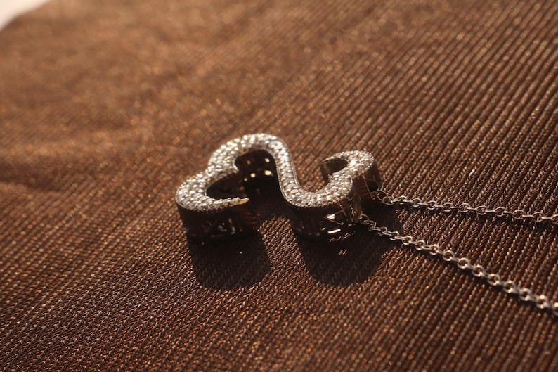 May include: A silver-colored pendant necklace featuring a double heart design. The pendant is encrusted with small, sparkling stones. The necklace chain is delicate and silver, resting on a brown textured surface.