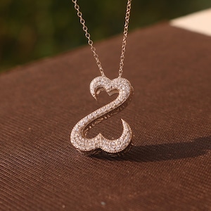 May include: A gold-colored pendant necklace featuring a double-heart design encrusted with small, sparkling stones. The pendant hangs from a delicate chain. The necklace is displayed on a brown surface.