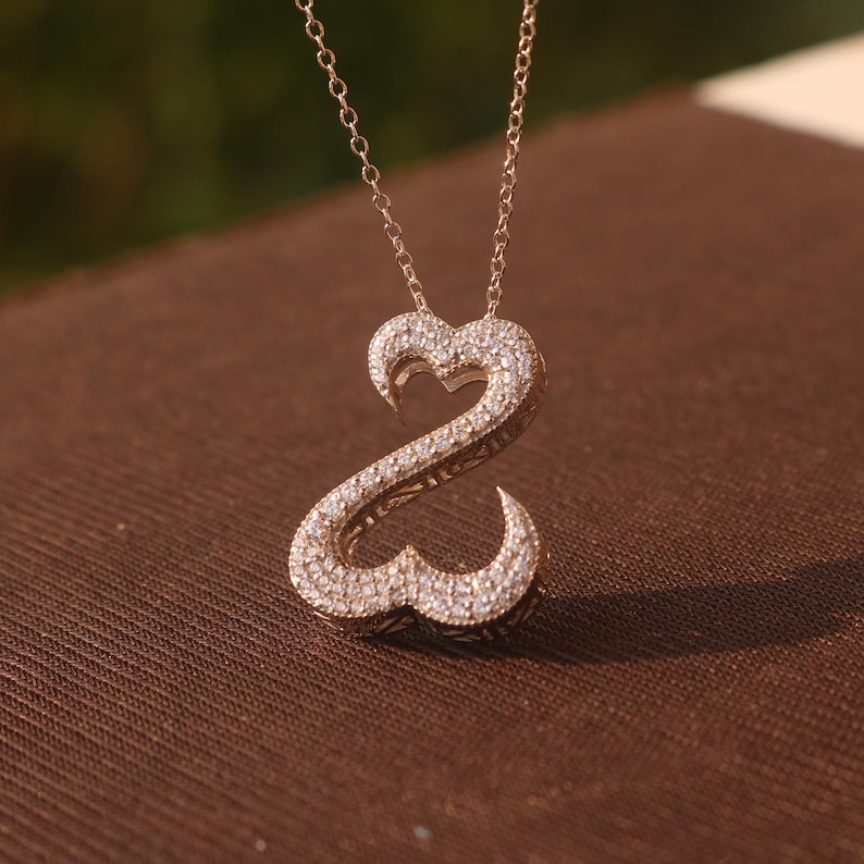 May include: A gold-colored pendant necklace featuring a double-heart design encrusted with small, sparkling stones. The pendant hangs from a delicate chain. The necklace is displayed on a brown surface.