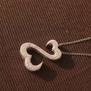 May include: A silver necklace with a pendant in the shape of an infinity symbol, encrusted with small, sparkling stones. The pendant is connected to a delicate chain, set against a textured brown background.