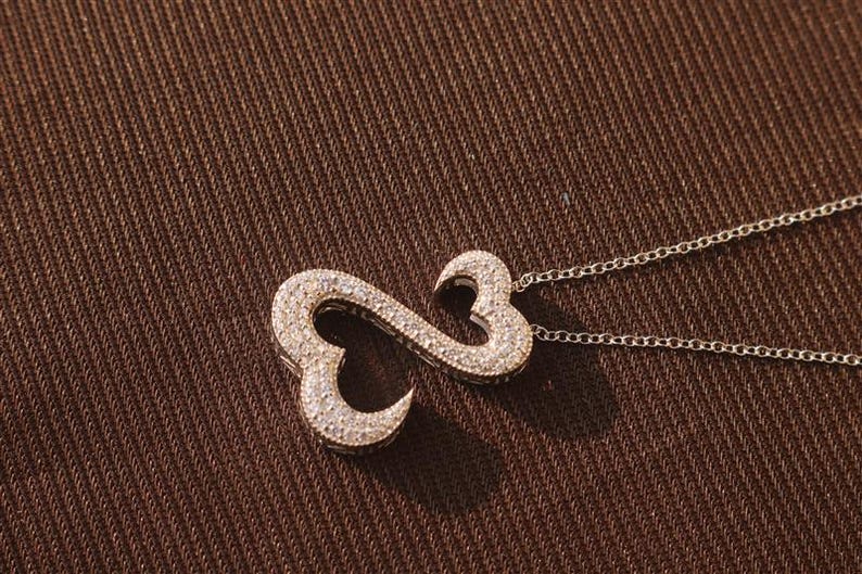 May include: A silver necklace with a pendant in the shape of an infinity symbol, encrusted with small, sparkling stones. The pendant is connected to a delicate chain, set against a textured brown background.