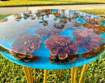 May include: Round side table with a clear blue resin top, featuring embedded brown pine cones. The table has bright yellow hairpin legs. The resin top reflects the surrounding trees and sky.