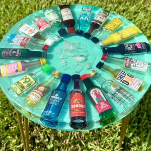 Może przedstawiać: Okrągły, turkusowy stół z żywicy ze złotymi nogami, prezentujący kolekcję miniaturowych butelek z alkoholem ułożonych w okrąg. Butelki mają różne marki i kolorowe etykiety, tworząc żywą ekspozycję. Stół stoi na zielonej trawie.