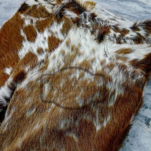 K&ouml;nnte beinhalten: Nahaufnahme von zwei St&uuml;cken Rindsleder. Das Leder hat ein nat&uuml;rliches Muster aus braunen und wei&szlig;en Flecken. Die Textur des Fells ist sichtbar, und die N&auml;hte sind entlang der Kanten sichtbar. Der Text "HAMI LEATHER" ist sichtbar.