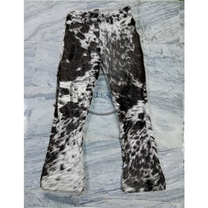 K&ouml;nnte beinhalten: Kuhfellhose mit einem schwarz-wei&szlig;en Fleckenmuster. Die Hose hat einen ausgestellten Bein-Design. Das Bild zeigt die Hose flach auf einer hellgrauen Oberfl&auml;che liegend. Die Hose ist aus einem weichen, strukturierten Material gefertigt.
