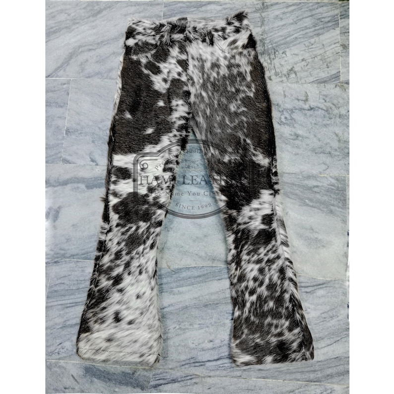 K&ouml;nnte beinhalten: Kuhfellhose mit einem schwarz-wei&szlig;en Fleckenmuster. Die Hose hat einen ausgestellten Bein-Design. Das Bild zeigt die Hose flach auf einer hellgrauen Oberfl&auml;che liegend. Die Hose ist aus einem weichen, strukturierten Material gefertigt.