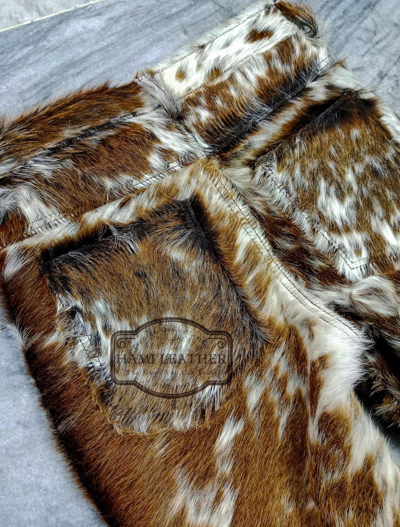 K&ouml;nnte beinhalten: Nahaufnahme einer Hose aus braunem, wei&szlig;em und schwarzem Kuhfell. Die Hose hat ein strukturiertes, geflecktes Muster und sichtbare N&auml;hte. Die Ges&auml;&szlig;tasche hat ein rechteckiges Etikett mit dem Text "HAMI LEATHER".