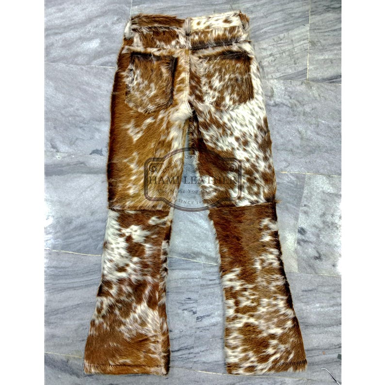 K&ouml;nnte beinhalten: Hose aus Kuhfell mit einem braun-wei&szlig; gefleckten Muster. Die Hose hat ein klassisches F&uuml;nf-Taschen-Design und ein ausgestelltes Bein. Das Kuhfellmaterial hat ein weiches, strukturiertes Aussehen. Die Hose liegt flach auf einer hellgrauen Oberfl&auml;che.