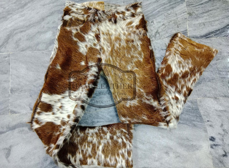 K&ouml;nnte beinhalten: Hose aus braun-wei&szlig;em Kuhfell. Die Hose hat ein nat&uuml;rliches Muster aus braunen und wei&szlig;en Flecken. Sie liegt flach auf einer grauen Oberfl&auml;che. Das Material scheint weich und strukturiert zu sein.