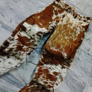 K&ouml;nnte beinhalten: Kuhfellhose in Braun- und Wei&szlig;t&ouml;nen. Die Hose besteht aus einem nat&uuml;rlichen Kuhfellmaterial mit einem einzigartigen Fleckenmuster. Die Hose liegt flach auf einer grauen Fliesenoberfl&auml;che. Das Logo "Hami Leather" ist sichtbar.