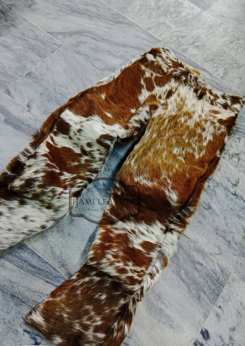 K&ouml;nnte beinhalten: Kuhfellhose in Braun- und Wei&szlig;t&ouml;nen. Die Hose besteht aus einem nat&uuml;rlichen Kuhfellmaterial mit einem einzigartigen Fleckenmuster. Die Hose liegt flach auf einer grauen Fliesenoberfl&auml;che. Das Logo "Hami Leather" ist sichtbar.