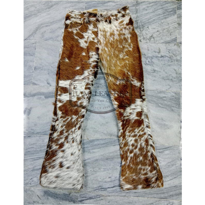 K&ouml;nnte beinhalten: Braune und wei&szlig;e Kuhfellhose mit ausgestelltem Bein. Das nat&uuml;rliche Muster aus braunen und wei&szlig;en Flecken ist sichtbar. Die Hose ist aus einem weichen, strapazierf&auml;higen Material gefertigt. Das Design erinnert an Westernmode.