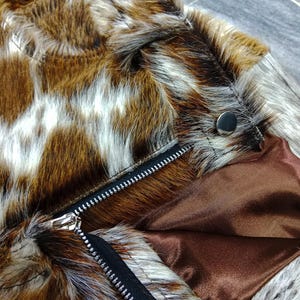 K&ouml;nnte beinhalten: Nahaufnahme einer Kunstfelljacke mit einem braunen, wei&szlig;en und beigefarbenen Muster. Die Jacke hat einen schwarzen Rei&szlig;verschluss und ein braunes Satinfutter. Ein silberner Druckknopf ist in der N&auml;he des Kragens zu sehen.