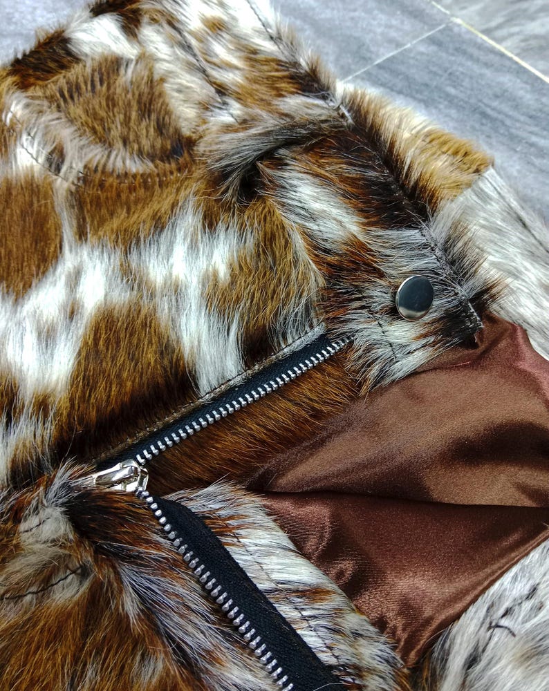 K&ouml;nnte beinhalten: Nahaufnahme einer Kunstfelljacke mit einem braunen, wei&szlig;en und beigefarbenen Muster. Die Jacke hat einen schwarzen Rei&szlig;verschluss und ein braunes Satinfutter. Ein silberner Druckknopf ist in der N&auml;he des Kragens zu sehen.