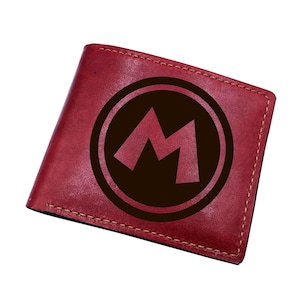 May include: A red bifold wallet featuring a dark brown circular emblem with a stylized letter M in the center. The wallet is constructed with visible stitching along the edges and has a smooth, matte texture throughout the exterior surface.