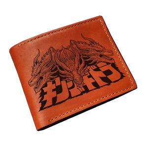 May include: A brown leather wallet featuring a detailed black illustration of a three-headed dragon and Japanese text. The wallet is rectangular and has a stitched border. The design is centered on the front of the wallet.