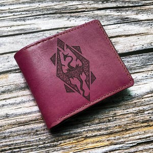 May include: A burgundy leather wallet featuring a black dragon emblem. The rectangular, folded wallet has stitched edges. The dragon design is centrally placed on the front. The background is a wooden surface.