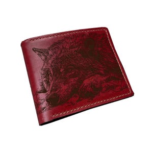 May include: A red leather wallet with a detailed engraving of a wolf's head. The wallet is square, with white stitching around the edges. The wolf's fur is intricately depicted, and the overall design is rustic.