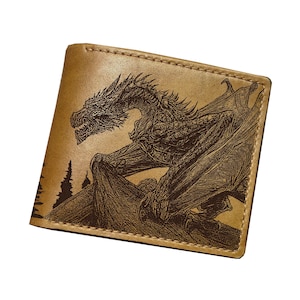 May include: A tan leather wallet featuring a detailed dragon engraving. The dragon is shown with its wings spread, and the design is etched into the leather. The wallet has a classic bi-fold design.